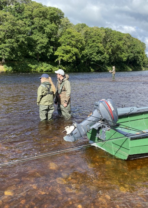 Here's some visiting salmon fishing guests receiving tuition behind the green painted moored up River Tay traditional salmon fishing boat. Look at the perfectly cut riverbank with the sun shining on it directly opposite these anglers.