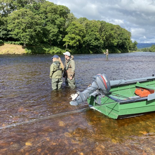 Here's some visiting salmon fishing guests receiving tuition behind the green painted moored up River Tay traditional salmon fishing boat. Look at the perfectly cut riverbank with the sun shining on it directly opposite these anglers.